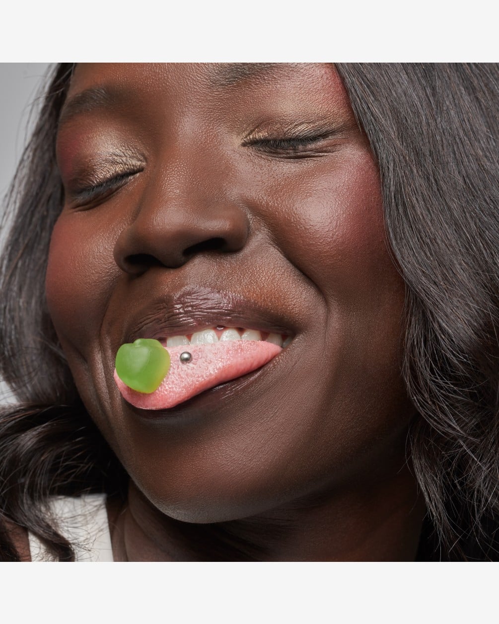 A close-up of a woman with dark wavy hair and a tongue piercing, smiling with her eyes closed as she holds a green SpoiledChild J29 gummy supplement on her tongue.