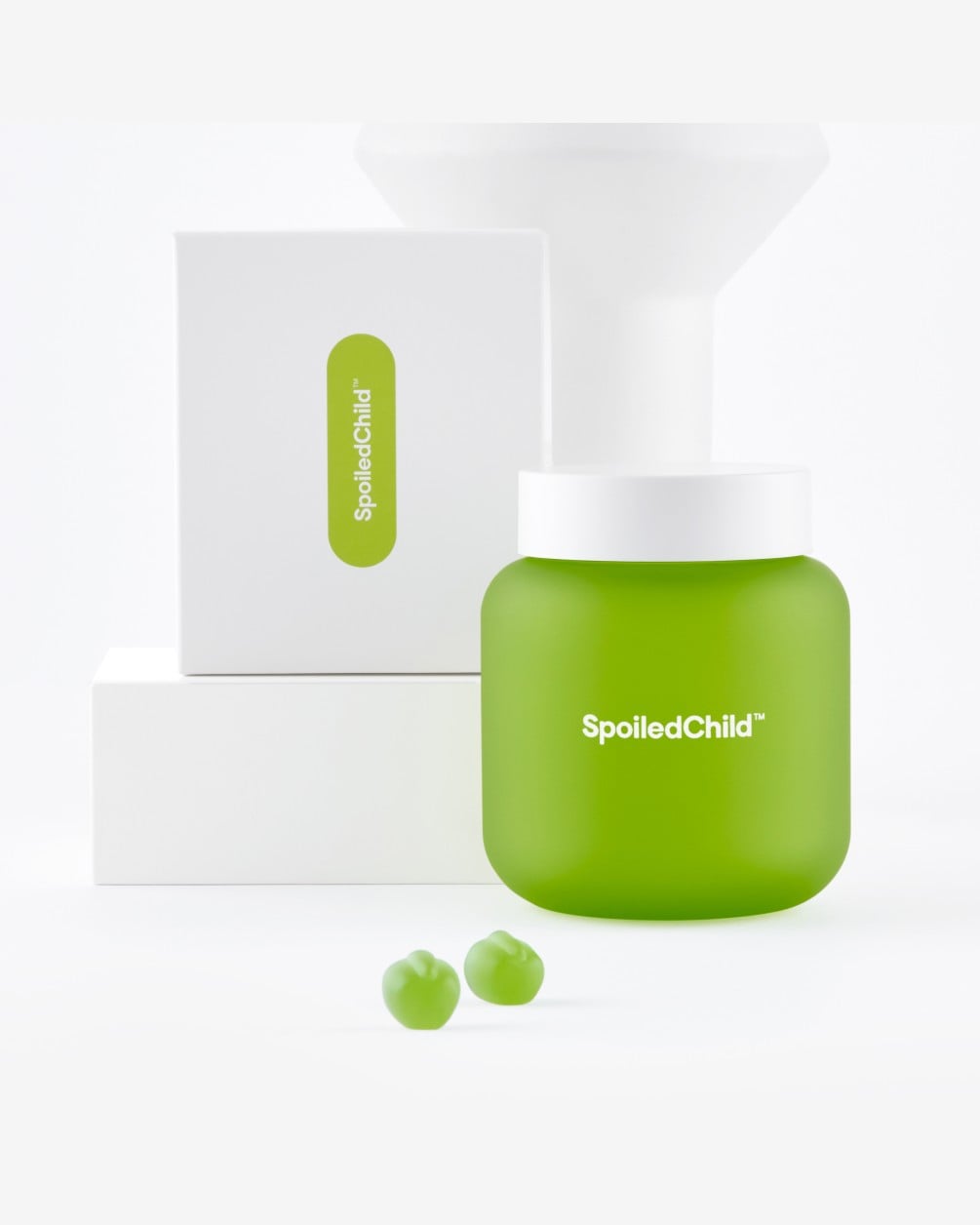 A green, square SpoiledChild J29 jar stands on a white surface next to two green gummy supplements. Behind the jar is its product box and a white sculptural vase.