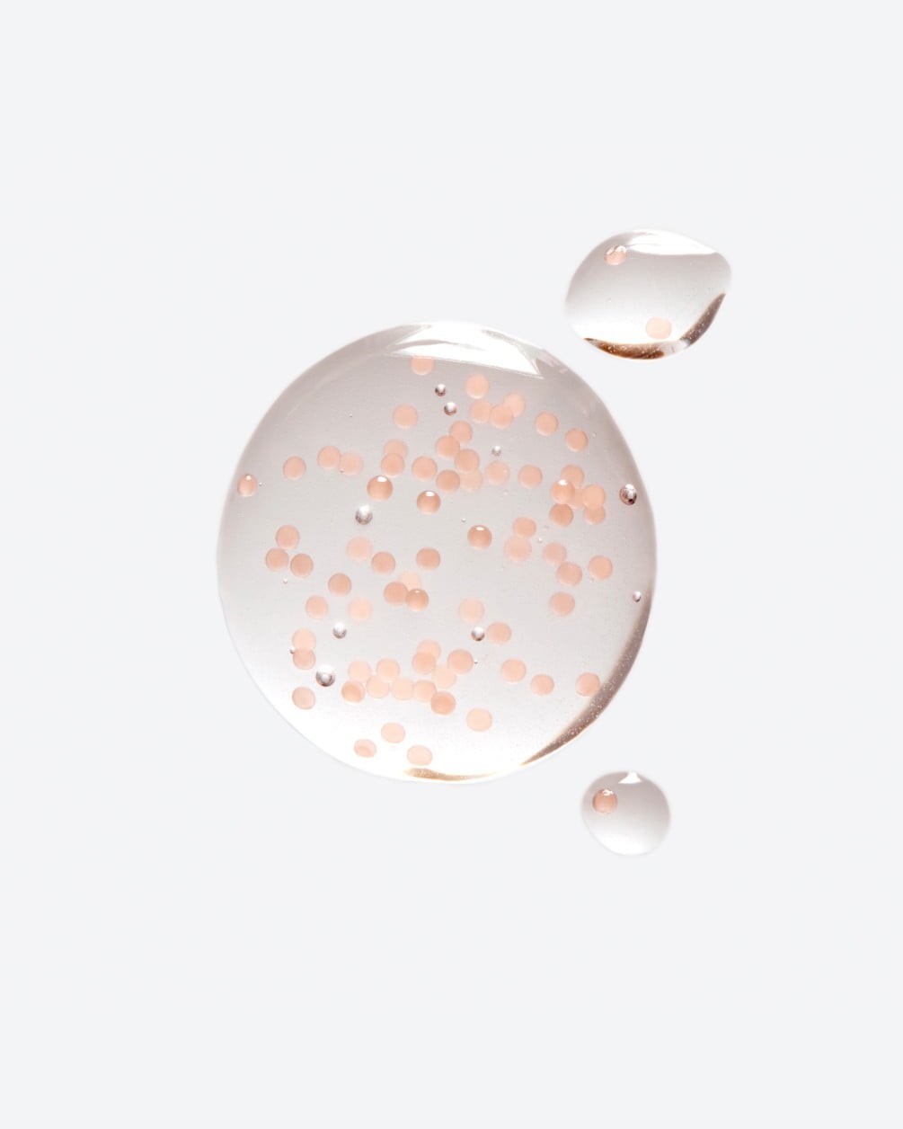 A close-up of several translucent, circular droplets of a liquid product on a white surface. The largest droplet contains numerous small, suspended pinkish-orange spheres or microbeads.